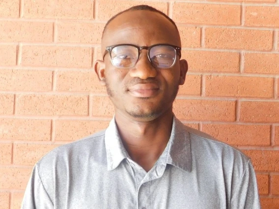 Prosper Babon-Ayeng - a man with very short hair, light facial hair, and square glasses standing against a brick wall. He is wearing a grey polo shirt. 