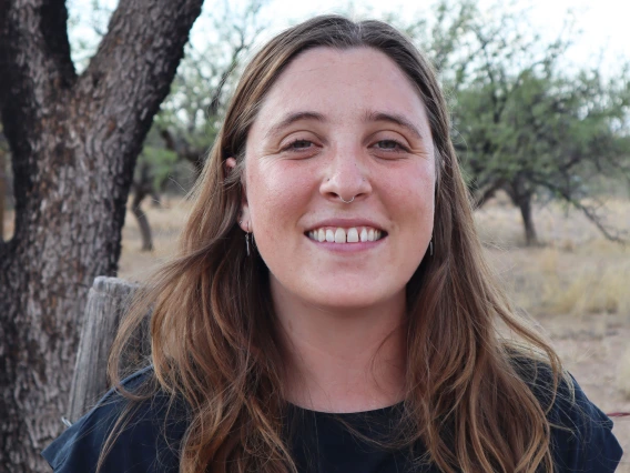 Madeleine Wallace - a woman with long brown hair wearing a blue shirt smiling at the camera and standing in front of a tree